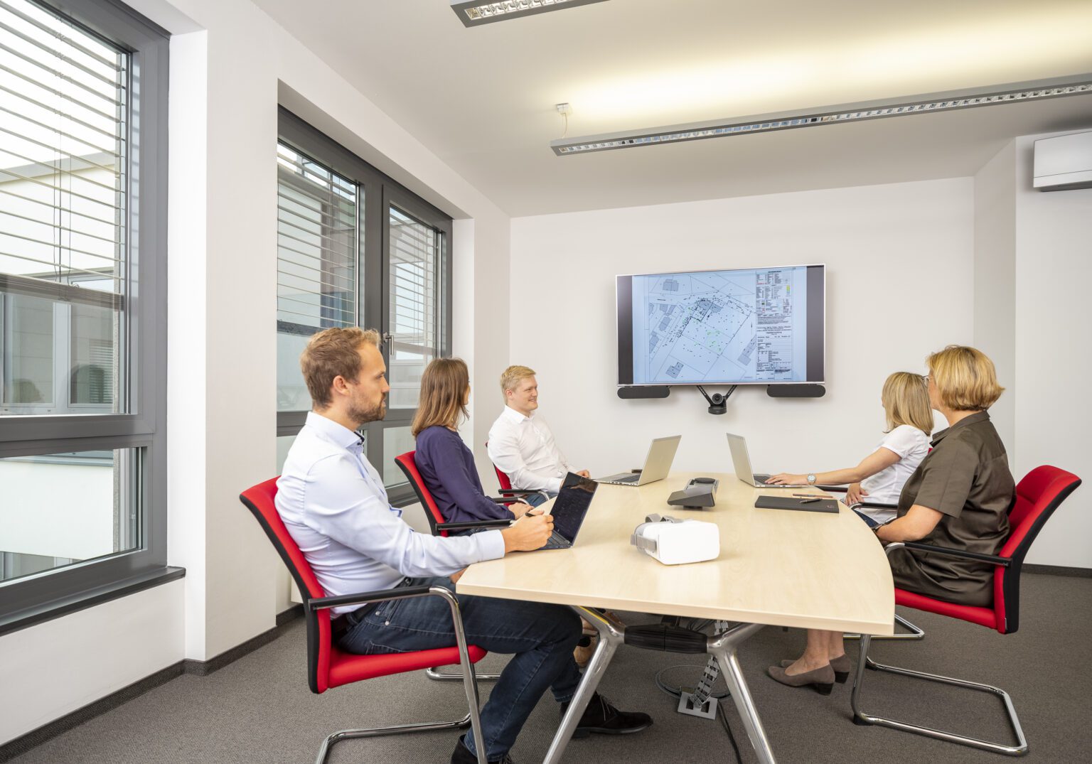 Meeting in modernem Konferenzraum, Personen mit Laptops diskutieren Baupläne auf Bildschirm, helle, professionelle Umgebung.