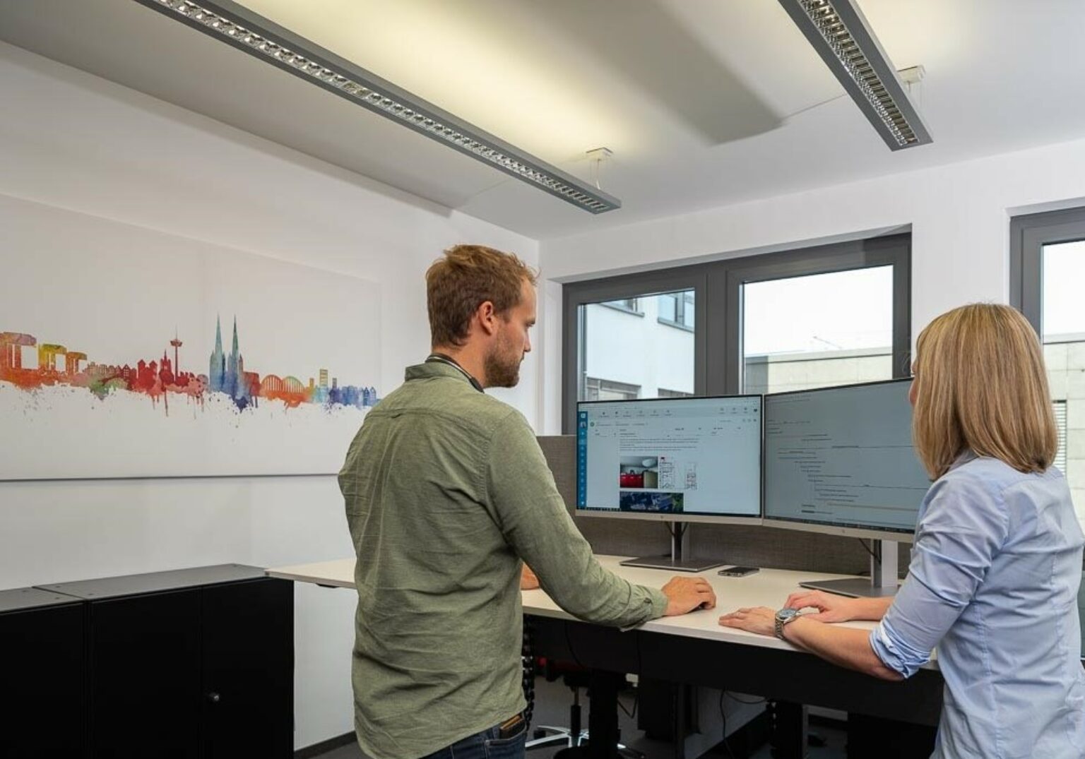 Zwei Personen arbeiten in einem modernen Büro an stehenden Schreibtischen mit doppelt gestapelten Monitoren.