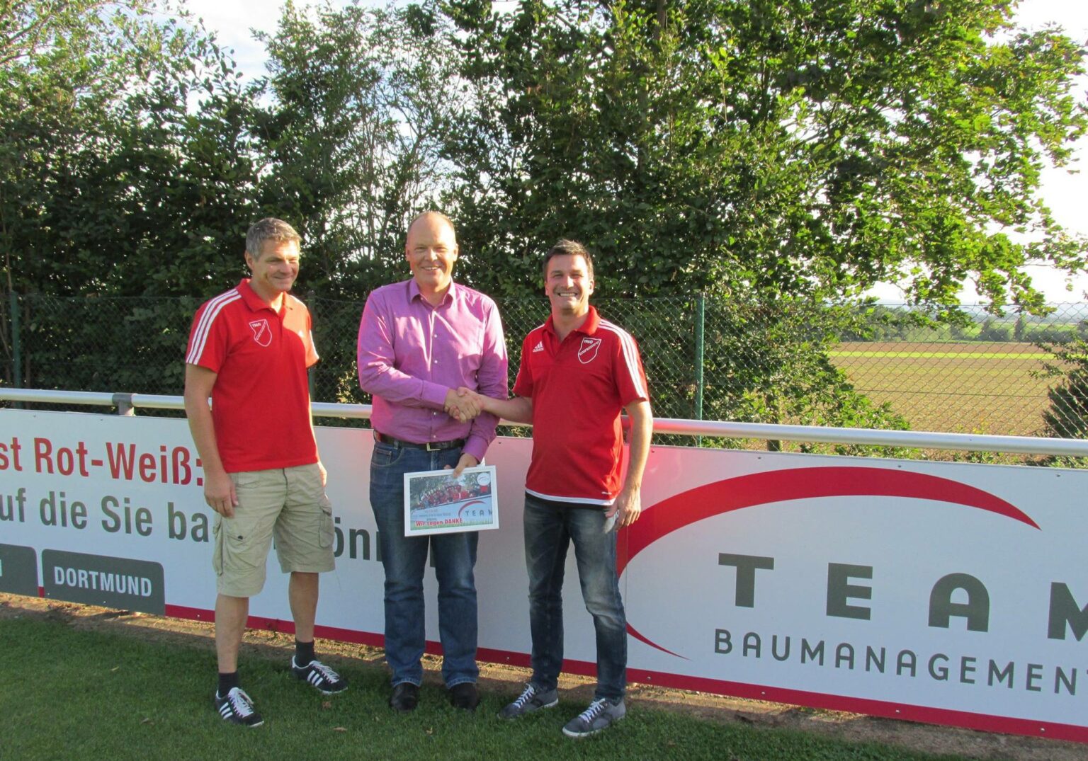 Drei Männer bei einer Händeschüttel-Zeremonie vor einem Werbeschild im Freien.