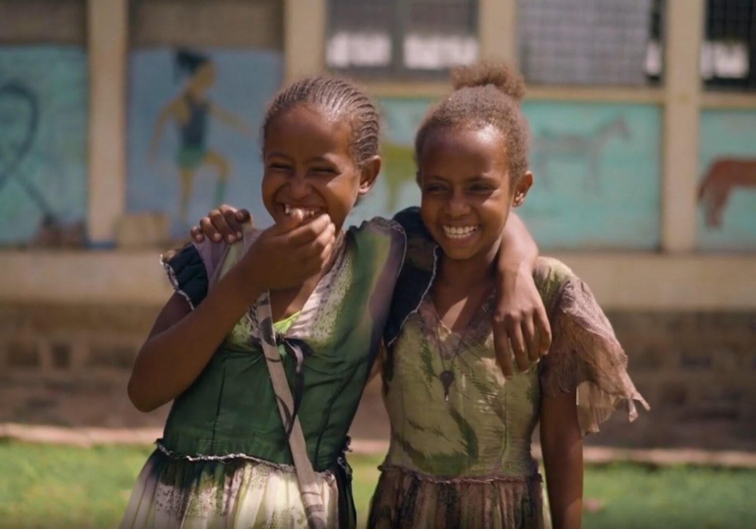 Lachende Mädchen umarmen sich im Schulhof, Hintergrund zeigt bunte Wandmalereien, fröhliche Stimmung.
