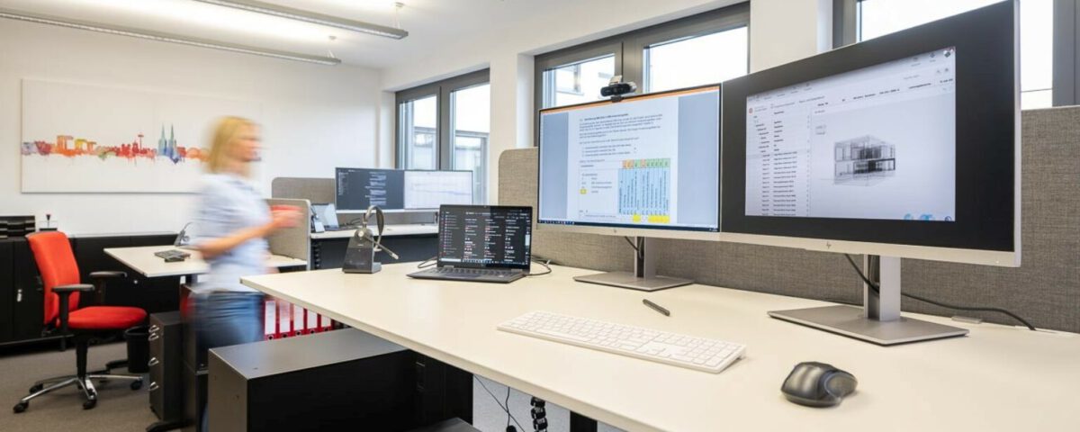 Modernes Büro mit höhenverstellbarem Schreibtisch, zwei Bildschirmen und Laptop. Person in Bewegung im Hintergrund.