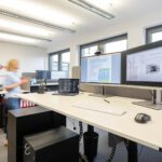 Modernes Büro mit höhenverstellbarem Schreibtisch, zwei Bildschirmen und Laptop. Person in Bewegung im Hintergrund.