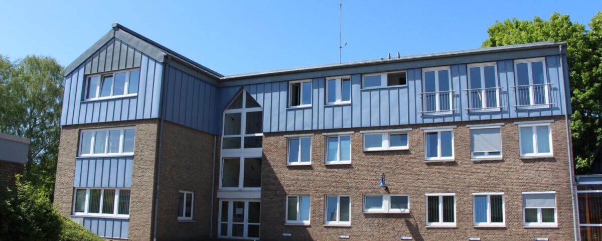 Moderner Bürogebäudekomplex aus Backstein mit blauen Paneelen an sonnigem Tag, umgeben von Natur.