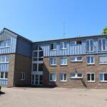 Moderner Bürogebäudekomplex aus Backstein mit blauen Paneelen an sonnigem Tag, umgeben von Natur.