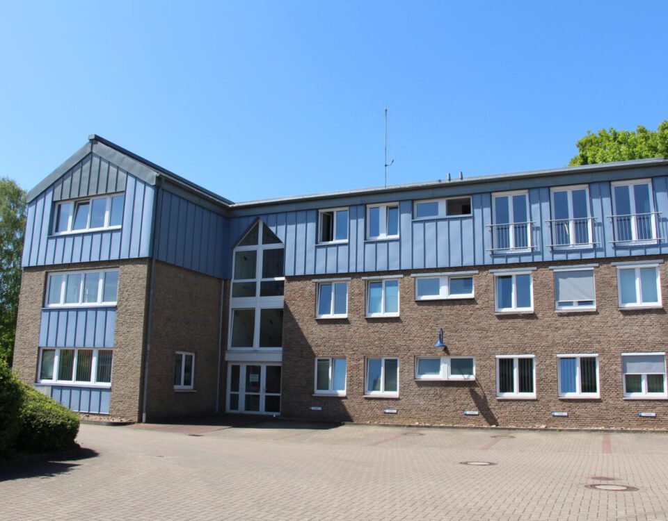 Moderner Bürogebäudekomplex aus Backstein mit blauen Paneelen an sonnigem Tag, umgeben von Natur.