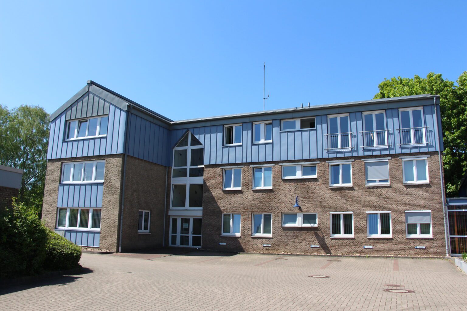 Moderner Bürogebäudekomplex aus Backstein mit blauen Paneelen an sonnigem Tag, umgeben von Natur.