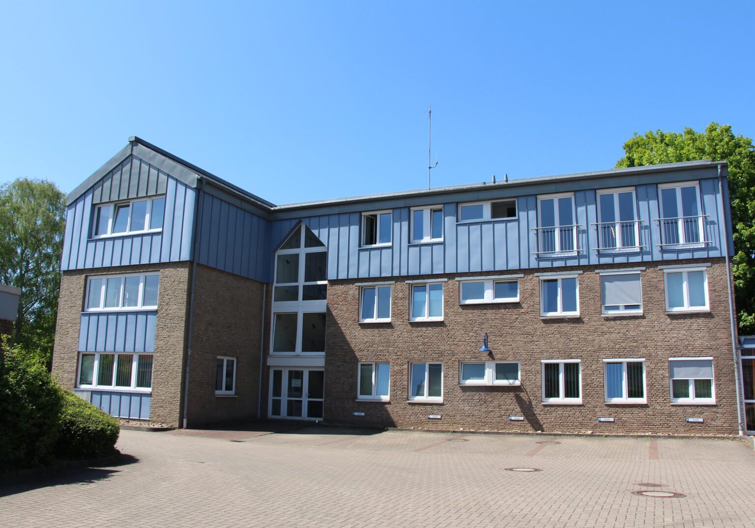 Backstein-Gebäude mit blauem Metalldach und großer Fensterfront bei sonnigem Wetter.