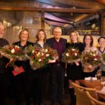 Gruppe von Personen mit Blumensträußen in einem festlichen Restaurant, freudig lächelnd für ein Gruppenfoto.