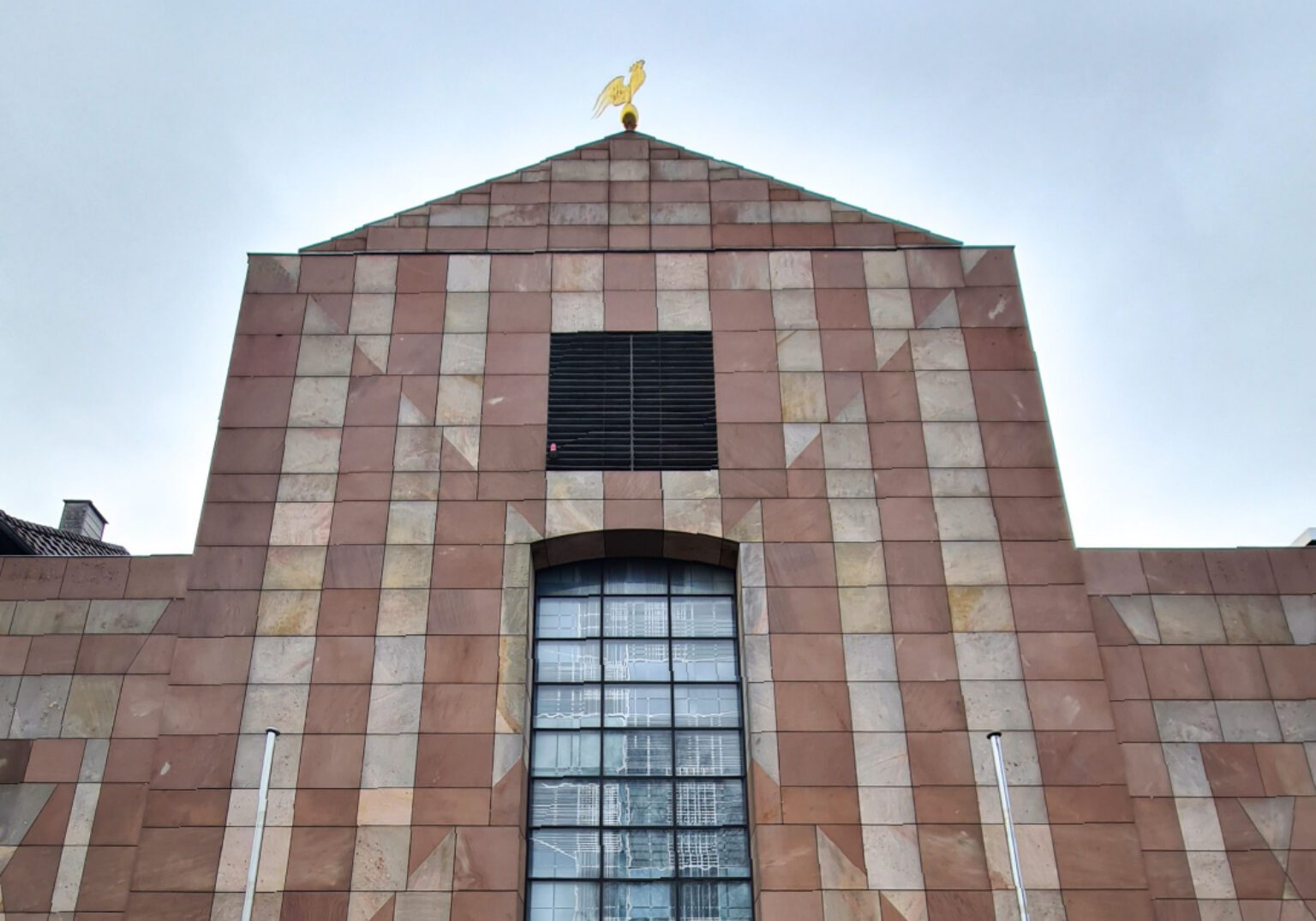 Gebäude mit markanter Fassadenstruktur und goldener Statue auf dem Dach unter bewölktem Himmel.