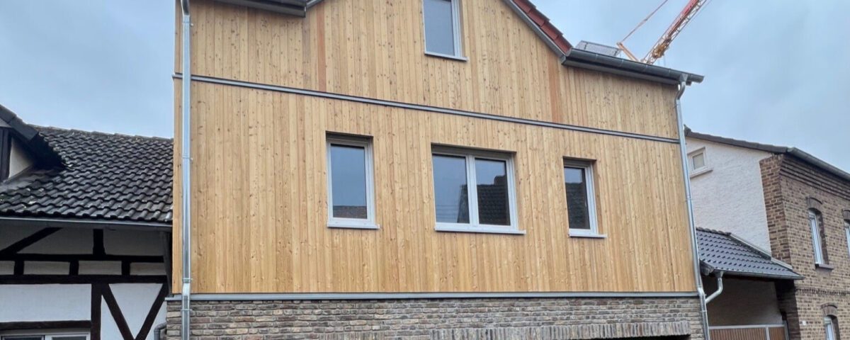 Modernes zweistöckiges Haus mit Holzfassade und grüner Garage in einer Wohngegend bei bewölktem Himmel.