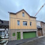Modernes zweistöckiges Haus mit Holzfassade und grüner Garage in einer Wohngegend bei bewölktem Himmel.