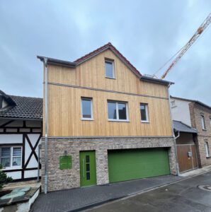 Modernes zweistöckiges Haus mit Holzfassade und grüner Garage in einer Wohngegend bei bewölktem Himmel.