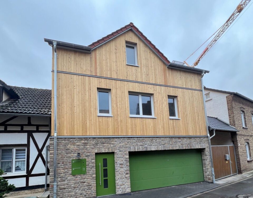 Modernes zweistöckiges Haus mit Holzfassade und grüner Garage in einer Wohngegend bei bewölktem Himmel.