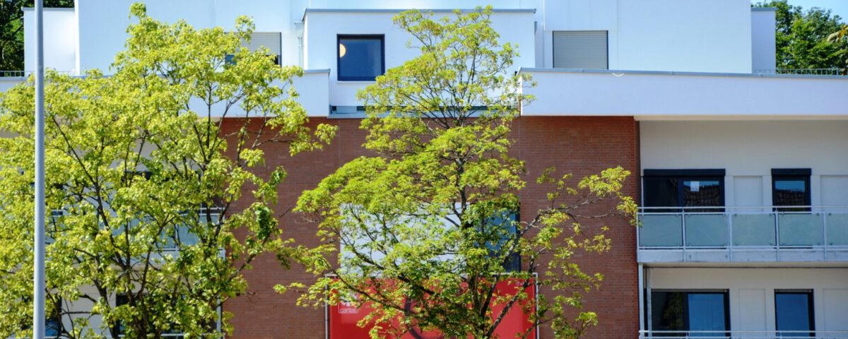 Mehrfamilienhaus aus Backstein mit Balkon und Bäumen, darunter ein Werbeplakat für Nächstenliebe von Caritas.