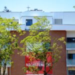 Mehrfamilienhaus aus Backstein mit Balkon und Bäumen, darunter ein Werbeplakat für Nächstenliebe von Caritas.