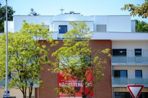 Mehrfamilienhaus aus Backstein mit Balkon und Bäumen, darunter ein Werbeplakat für Nächstenliebe von Caritas.