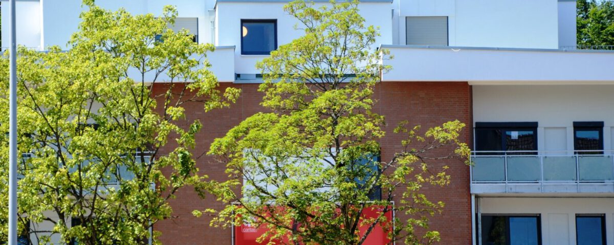 Moderner Wohnblock mit rotem Plakat und Bäumen im Vordergrund bei sonnigem Wetter, verdeutlicht Stadtleben und Architektur.