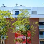 Moderner Wohnblock mit rotem Plakat und Bäumen im Vordergrund bei sonnigem Wetter, verdeutlicht Stadtleben und Architektur.