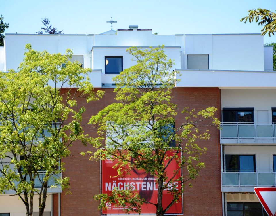 Moderner Wohnblock mit rotem Plakat und Bäumen im Vordergrund bei sonnigem Wetter, verdeutlicht Stadtleben und Architektur.