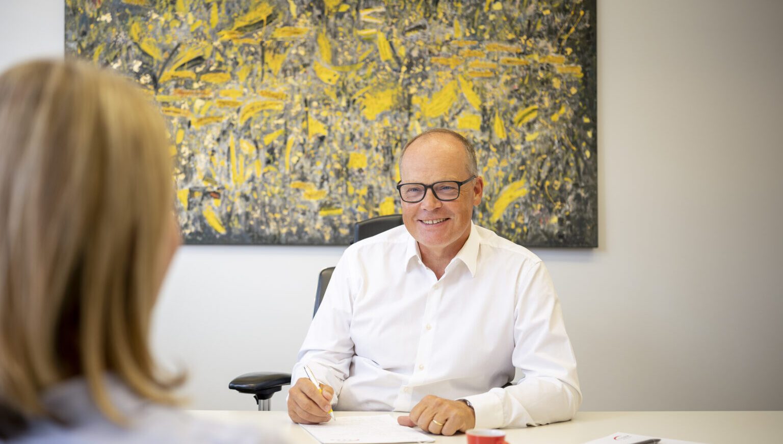 2311_064_AUSWAHL_18-Motiv_Externe-Kommunikation-6x7-1-1536x1075.2-c-default Geschäftsmann im weißen Hemd lächelt bei einem Gespräch in seinem Büro mit abstraktem Kunstwerk im Hintergrund.