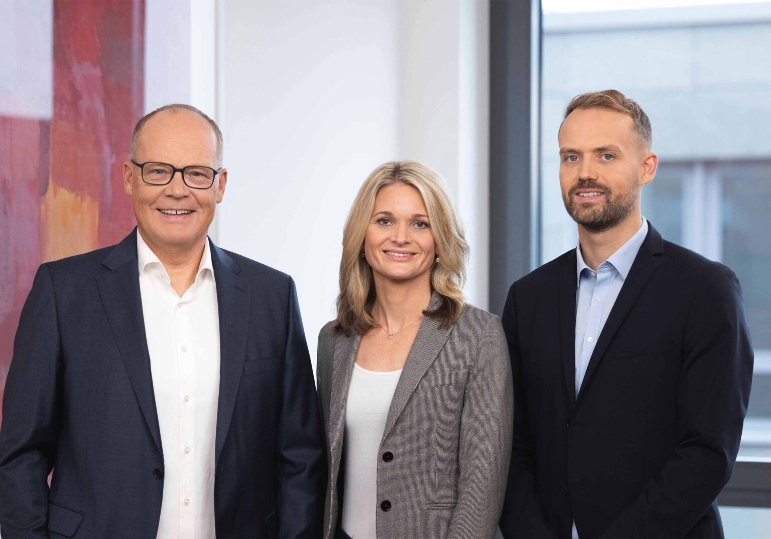 Drei Geschäftsleute in Anzügen stehen lächelnd in einem modernen Büro vor einem Fenster.