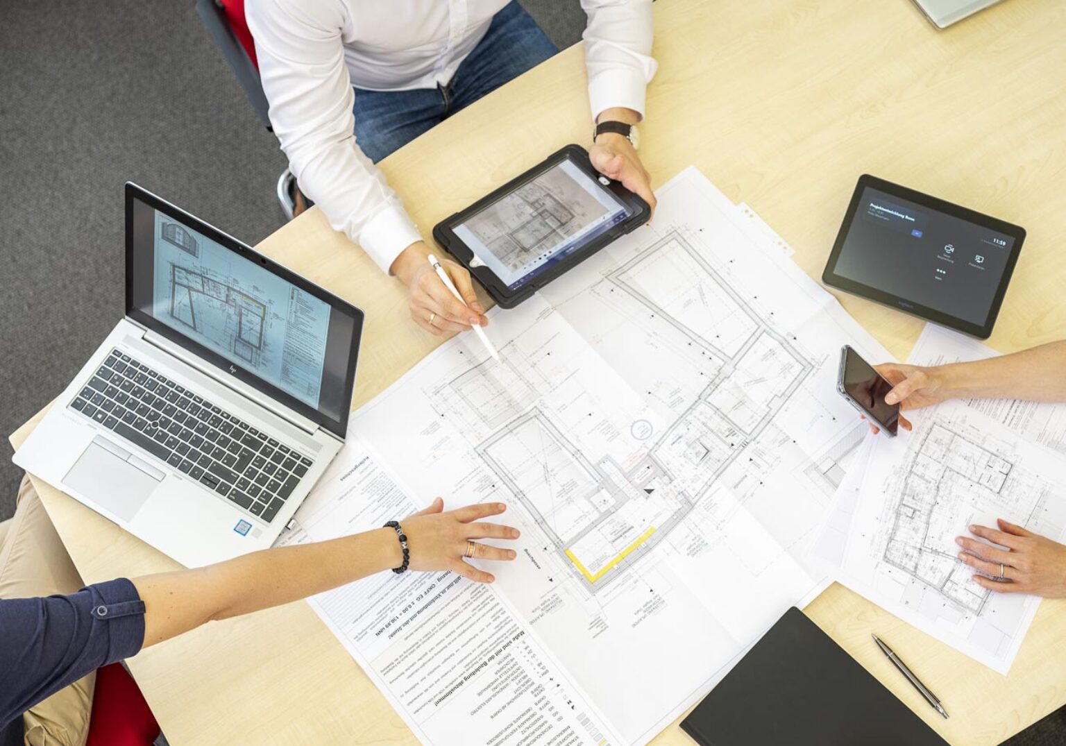 Architekten-Team diskutiert Baupläne am Tisch mit Laptop und Tablets.