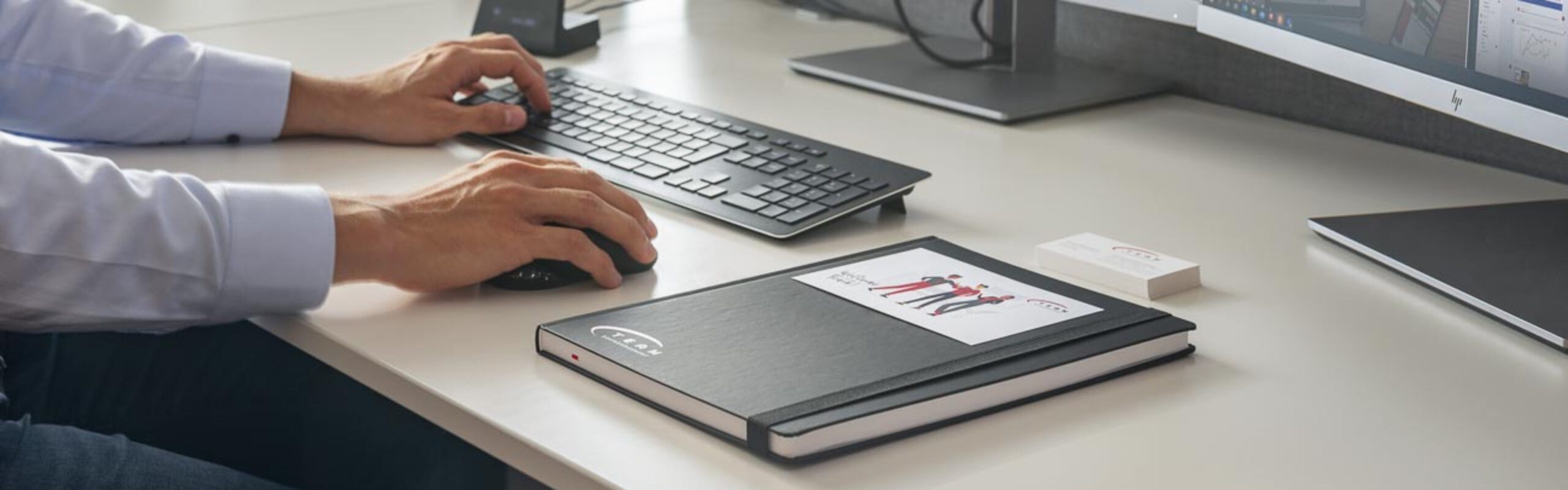 Büroarbeitsplatz mit Computer, Tastatur und Notizbuch auf einem Schreibtisch, Person tippt und nutzt Maus.