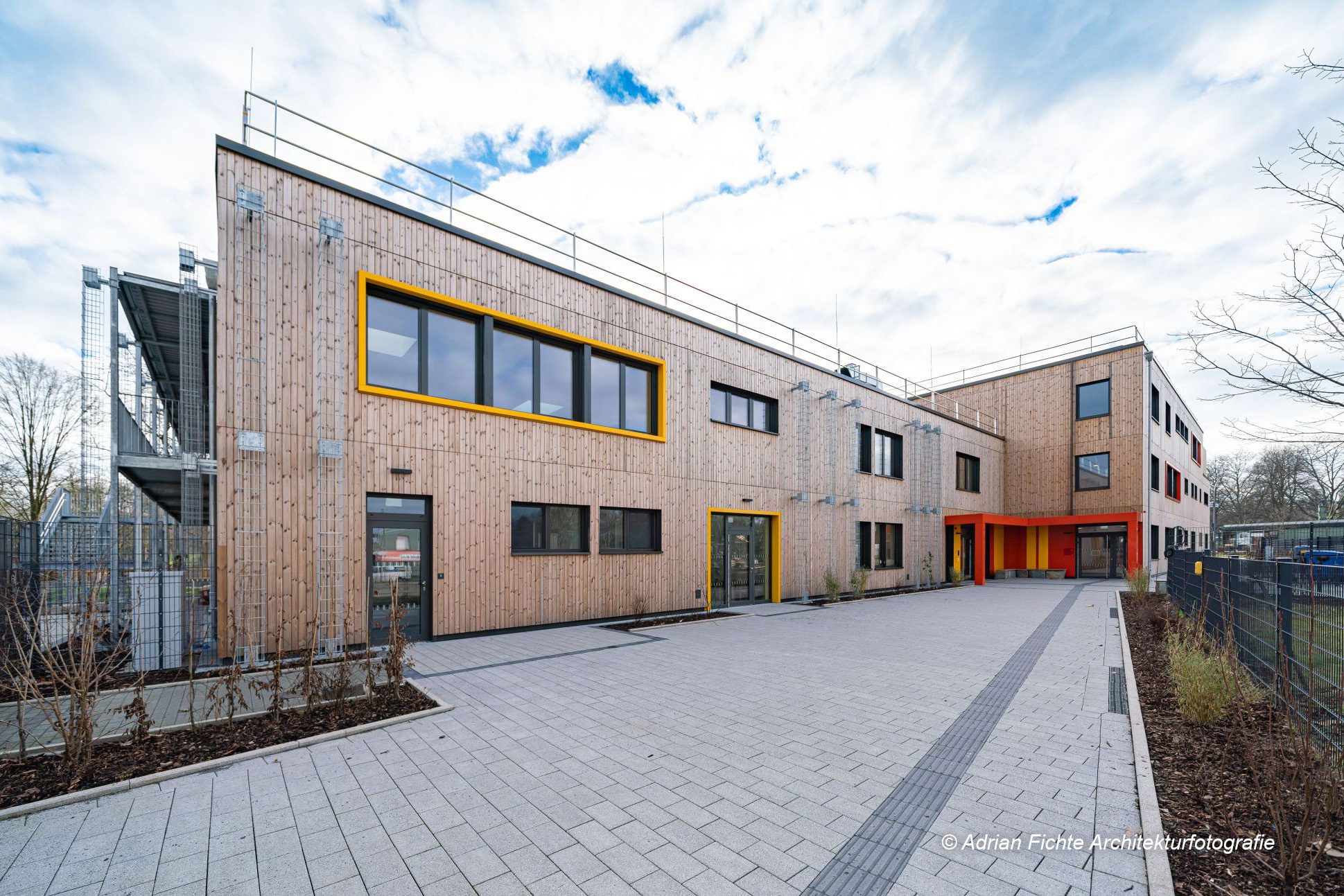 Modernes Gebäude mit Holzfassade und gelben Akzenten bei bedecktem Himmel, umgeben von einem gepflasterten Weg.