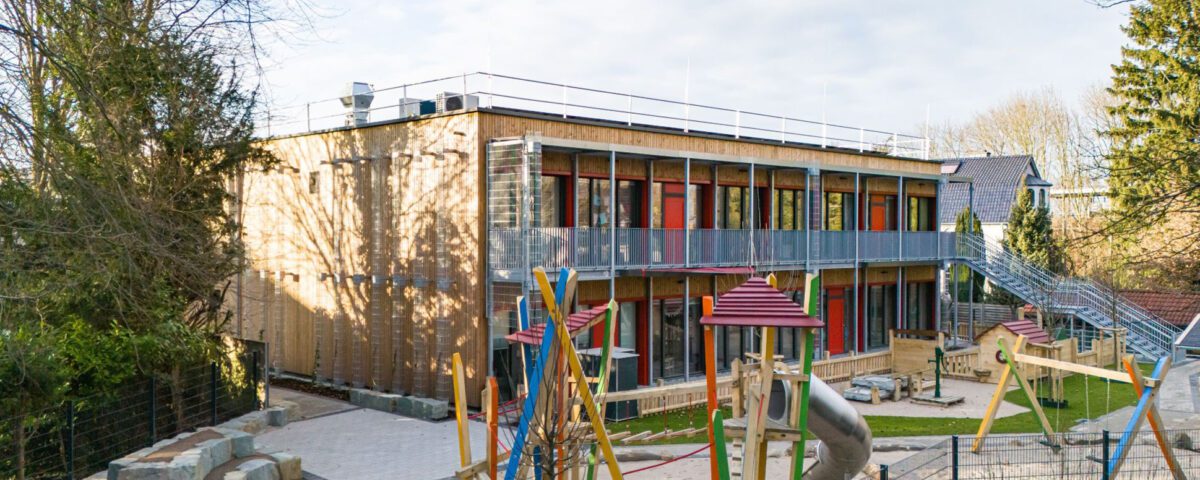 Modernes zweistöckiges Kindergarten-Gebäude mit buntem Spielplatz im Vordergrund, von Bäumen umgeben unter blauem Himmel.