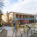 Modernes zweistöckiges Kindergarten-Gebäude mit buntem Spielplatz im Vordergrund, von Bäumen umgeben unter blauem Himmel.