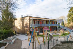 Modernes zweistöckiges Kindergarten-Gebäude mit buntem Spielplatz im Vordergrund, von Bäumen umgeben unter blauem Himmel.