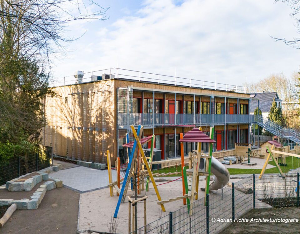 Modernes zweistöckiges Kindergarten-Gebäude mit buntem Spielplatz im Vordergrund, von Bäumen umgeben unter blauem Himmel.