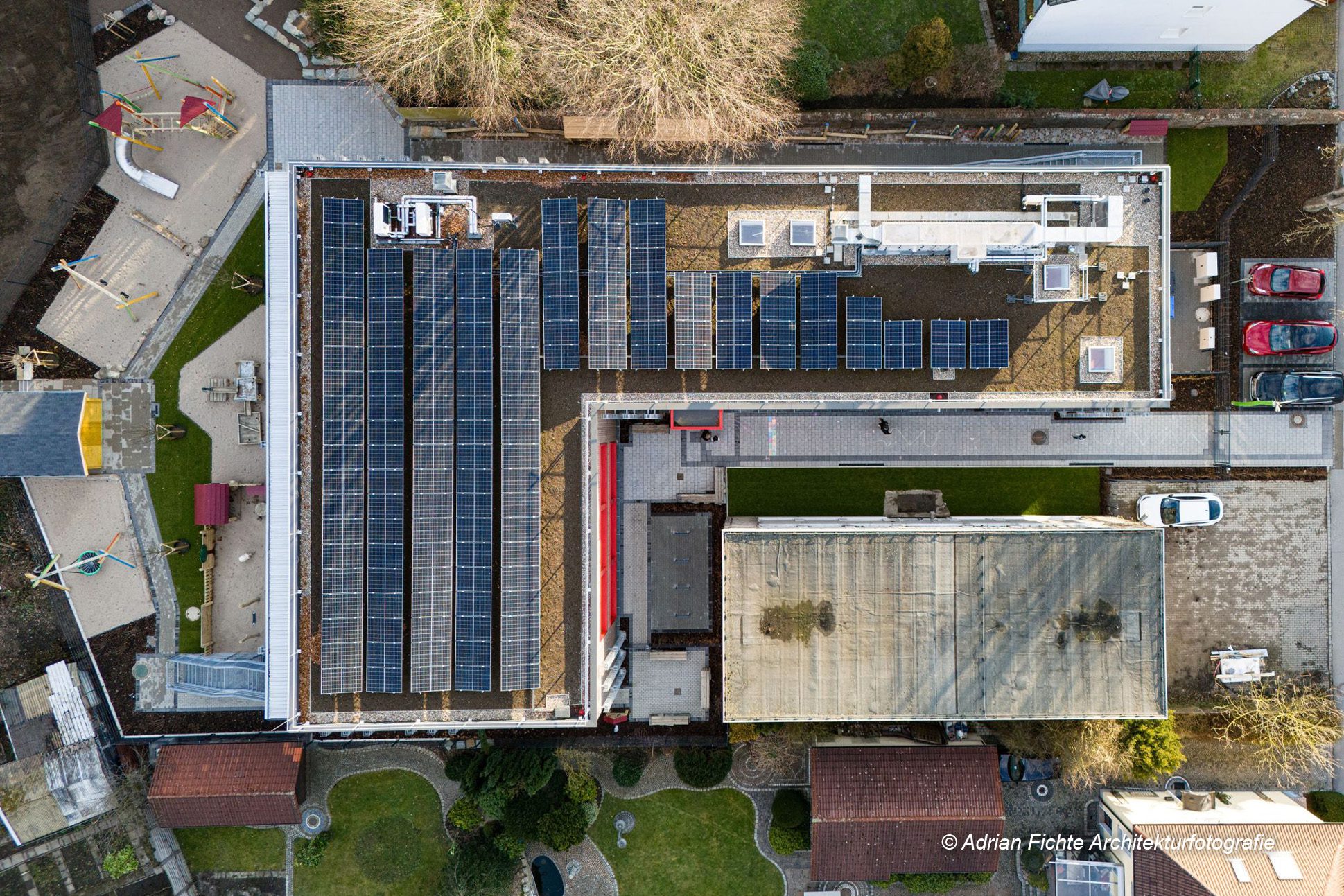 Dach eines Gebäudes mit Solaranlagen, umgeben von Parkplätzen und Grünflächen, Luftansicht.
